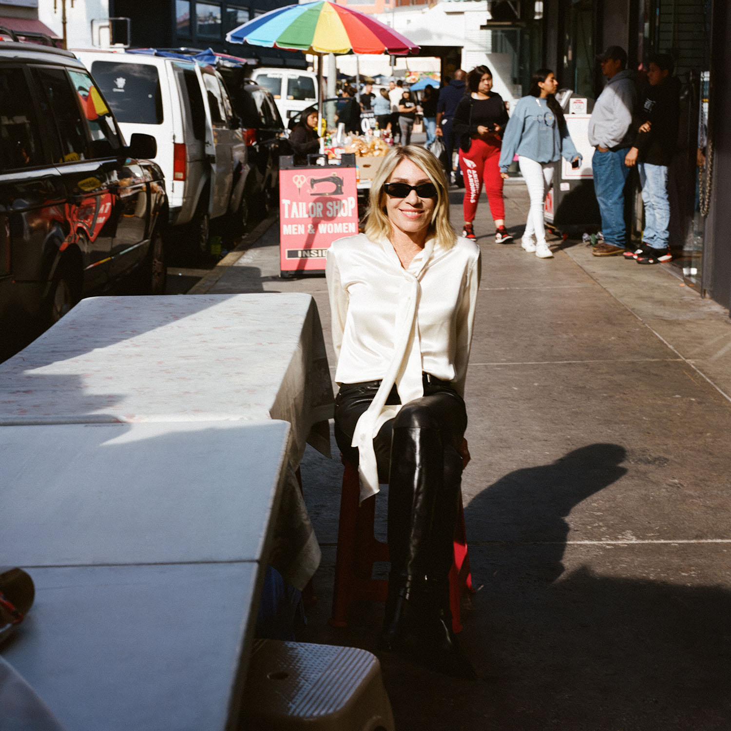 Color photograph of Kim Gordon sitting and smiling on the sidewalk of a street in New York City, by Danielle Neu